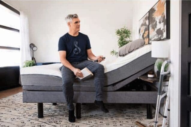 A man with gray hair sits on the edge of a hospital bed, holding a remote and a book. The adjustable bed is in a partially raised position. The room has light walls, a patterned rug, and a small plant on a nightstand.